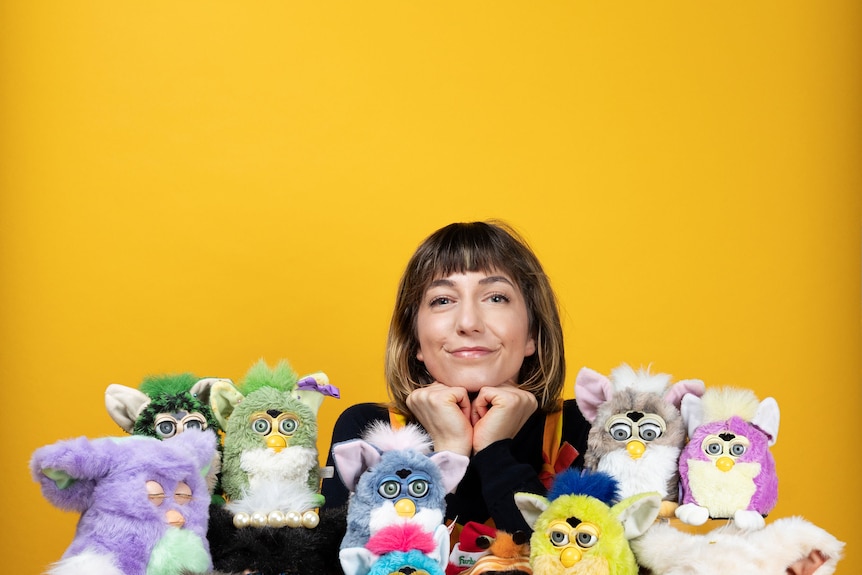 A portrait of a woman against a bright yellow background with a row of Furbies in front of her.