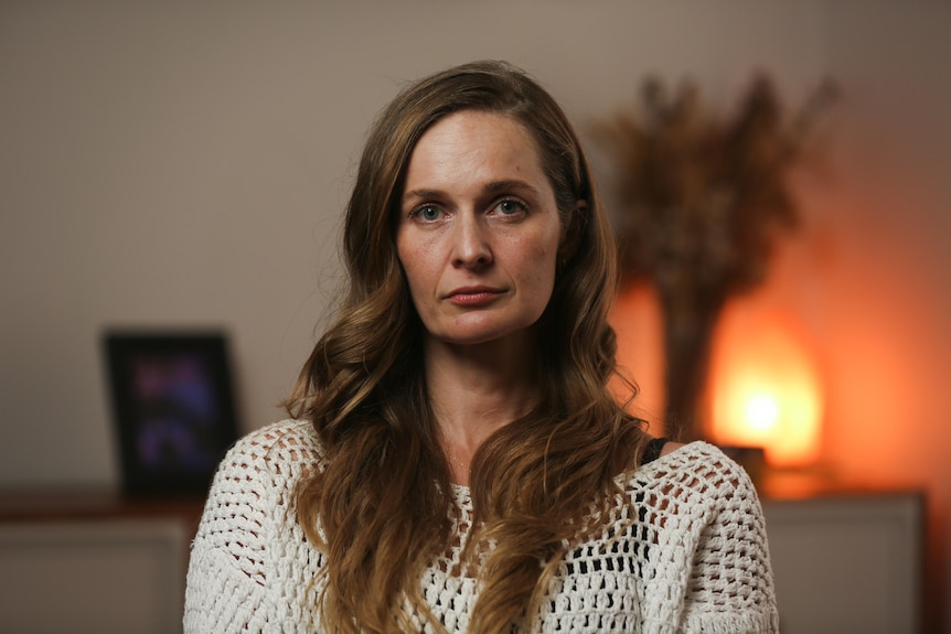 A woman sitting indoors looks into the camera with a neutral expression. Behind her is a shelf with flowers and a lamp.