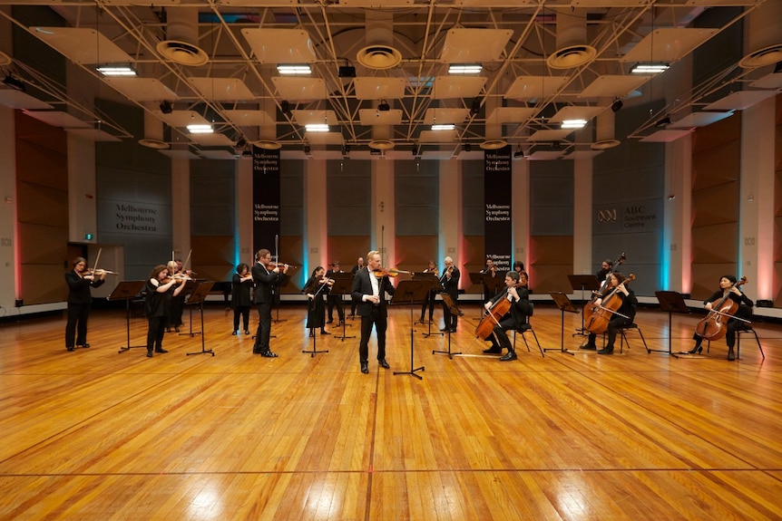 An orchestra plays in a spacious timber-floored room.
