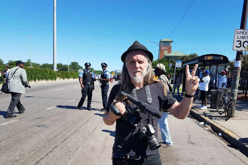 George Gittoes gives a peace sign while holding a video camera.