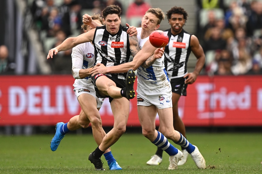 A Collingwood AFL player kicks the ball while being tackled by a North Melbourne opponent.