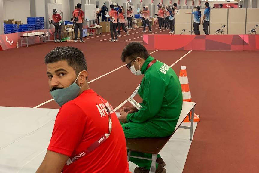 Two middle eastern men, one in red and the other in green, sit on a seat with face masks at indoor sports venue.