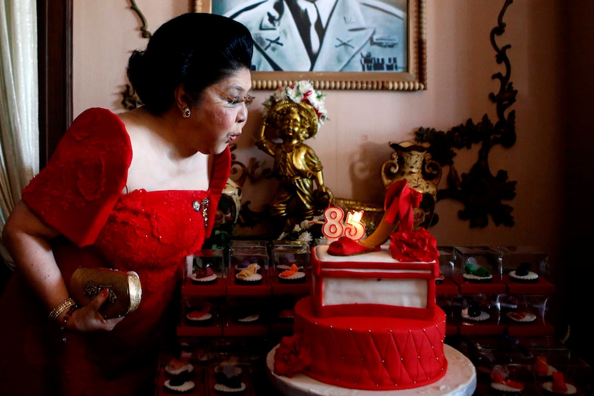 An older woman in a red dress blows out a candle on a red cake adorned with high-heeled shoes.