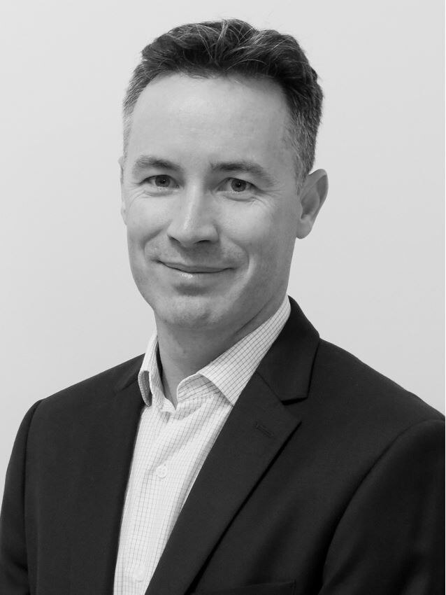 Black and white image of Peter McEvoy from chest up, wearing jacket and shirt, and smiling with mouth closed.