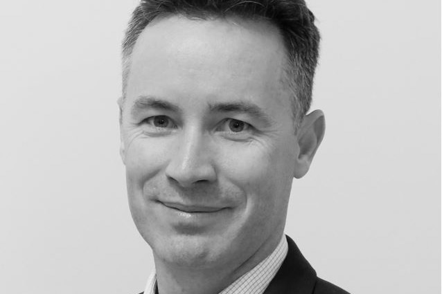 Black and white image of Peter McEvoy from chest up, wearing jacket and shirt, and smiling with mouth closed.