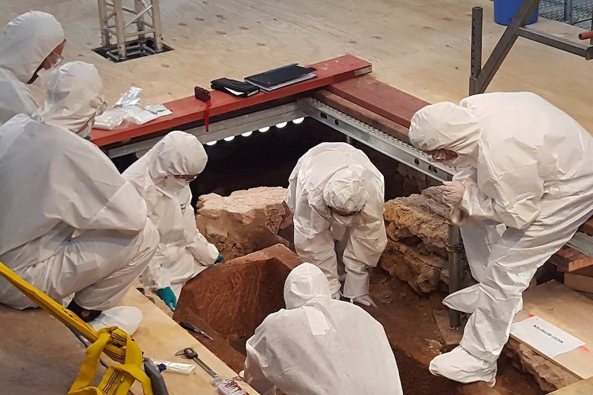 A group of people in protective gear look into a hole in a stone floor.