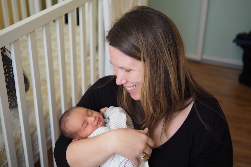 A woman with brown hair smiles as she looks down at a baby sleeping in her arms.
