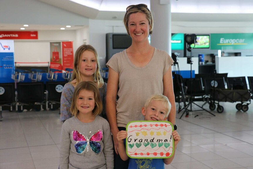 Melanie in an airport terminal with her two daughters and son, holding a sign that reads "grandma".