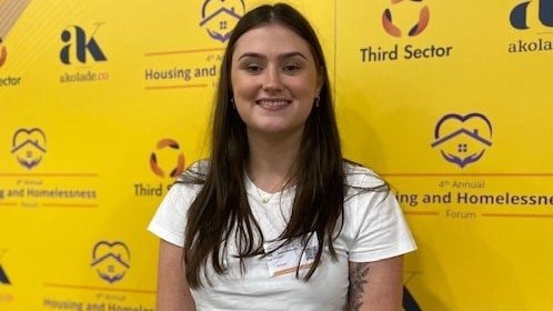 woman in white top with long brown hair stands in front of yellow poster smiling