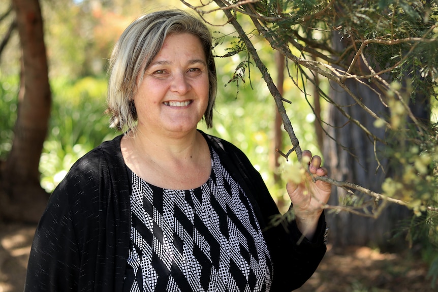 Kylie Ware wears a black cardigan over a black and white dress and holds a twig with some wattle flowers on it.