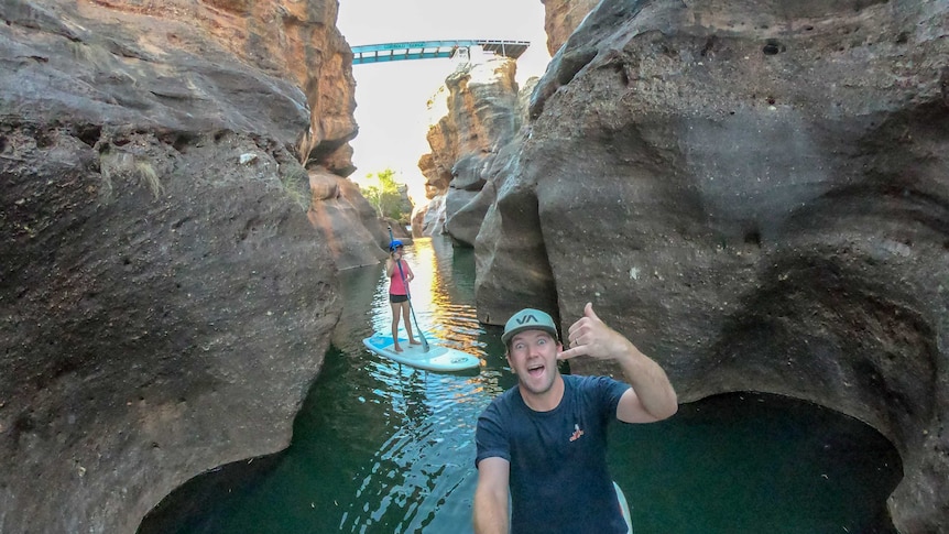 A man and woman stand-up paddle board through a gorge, with a bridge in the background.