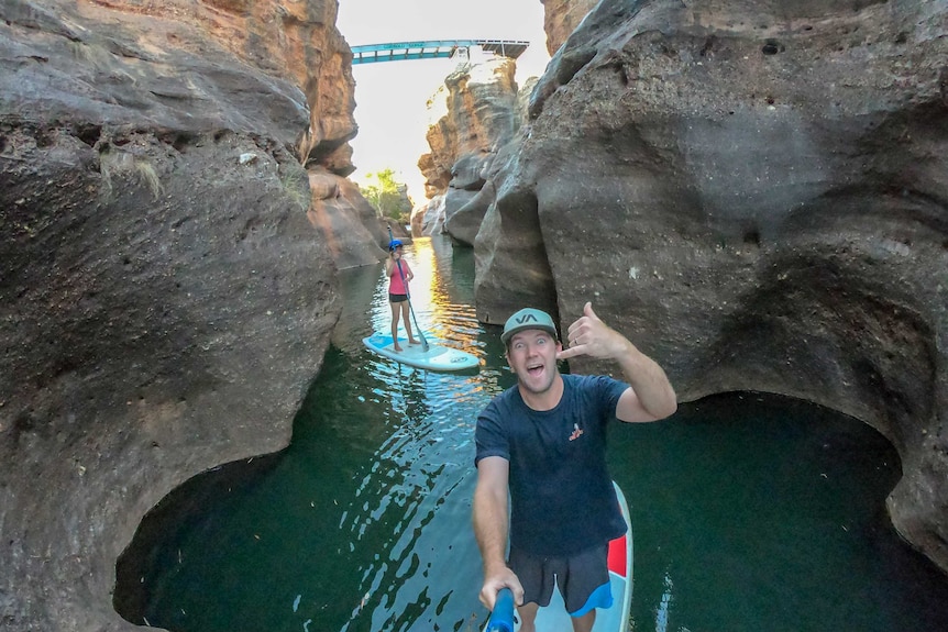 A man and woman stand-up paddle board through a gorge, with a bridge in the background.