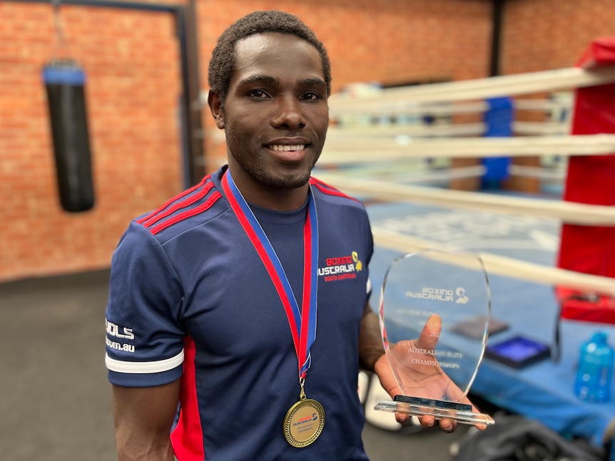 A young man who is wearing a gold medal around his neck smiles as he holds a glass trophy, a boxing ring behind him