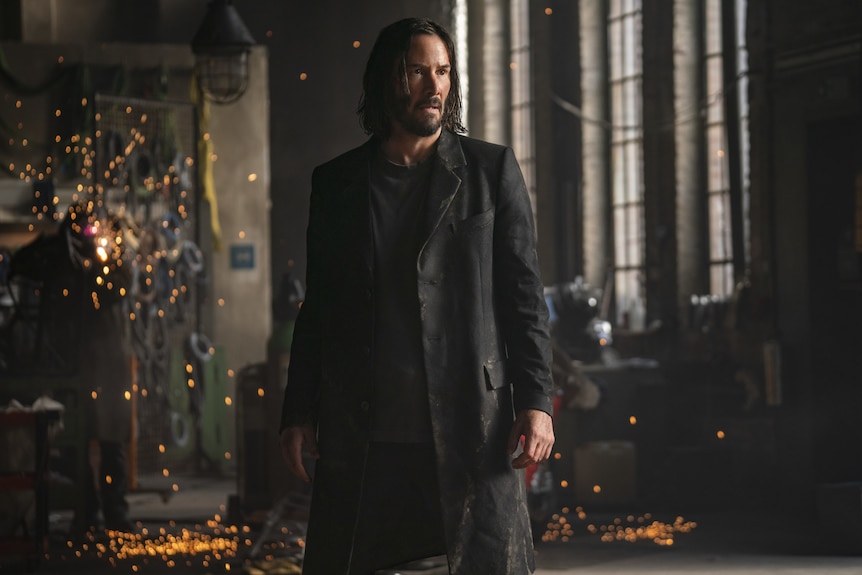 Un homme de 57 ans avec de longs cheveux noirs et une barbe dans un long manteau sombre se tient dans un couloir, des étincelles derrière lui