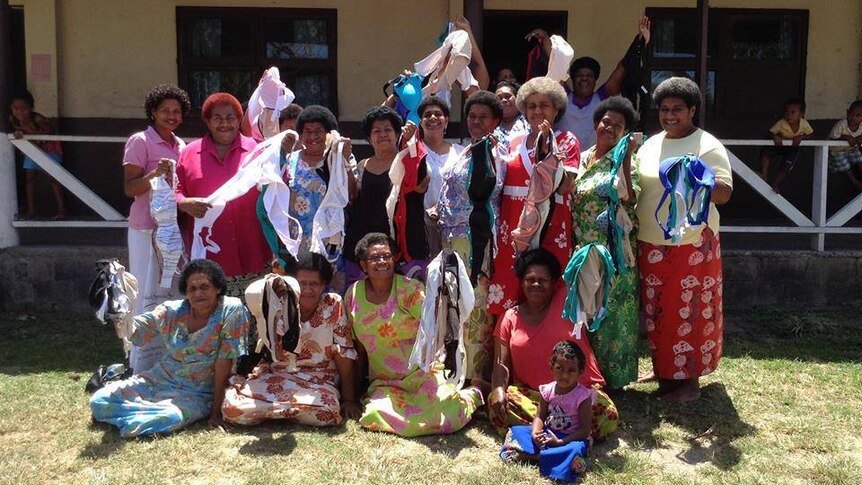 A group of Fijian women stand in front of a building and hold bras in the air.