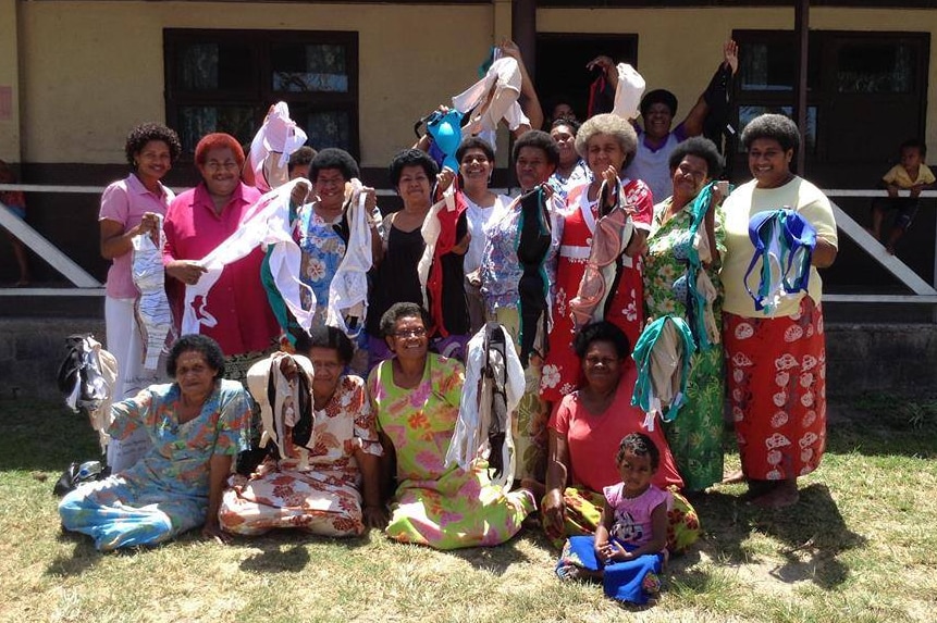 A group of Fijian women stand in front of a building and hold bras in the air.