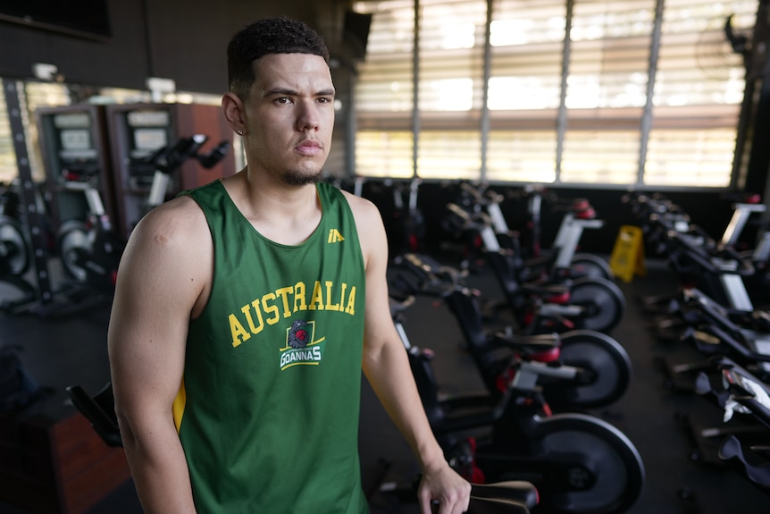Jarrod is wearing a dark green Australia jersey, looking pensive as he stands in a room full of exercise bikes.