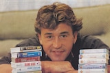 A man folds his arms on top of a table and positions his head behind cassette tapes stacked in towers.