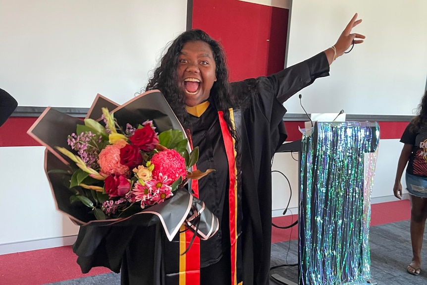 A woman with long dark hair wears a black graduation gown and holds a bunch of flowers. She looks very excited