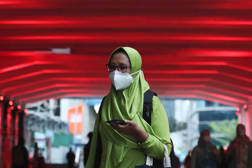 A women wears a face mask and a lime green hijab while holing a smartphone with red lights in the background.