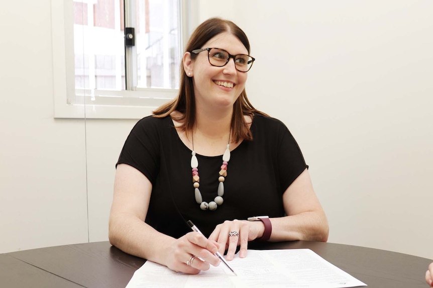 Sarah Dale est assise à un bureau avec des papiers devant elle.  Elle regarde et sourit à quelqu'un hors du cadre.