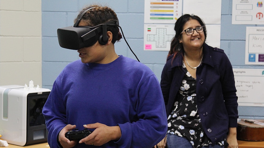 A girl in a blue sweatshirt holds a gaming controller while wearing black virtual reality goggles.