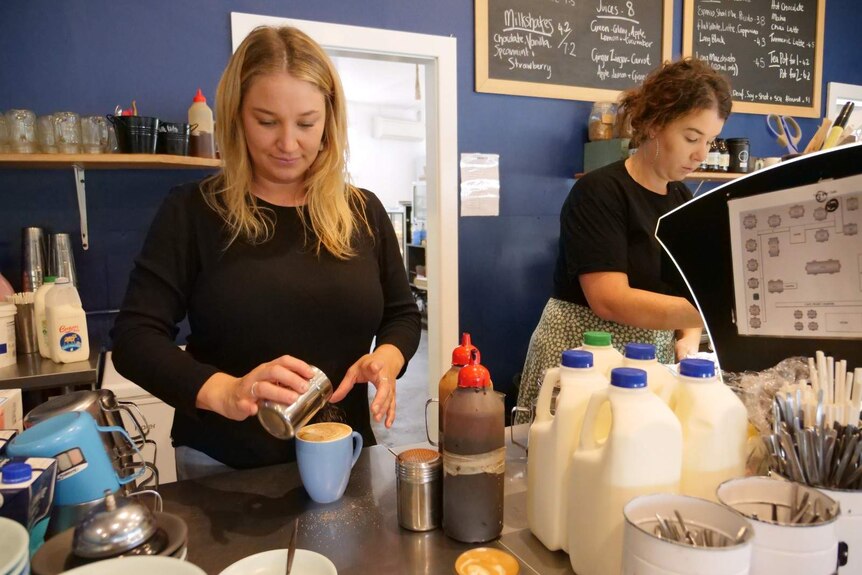 One women putting chocolate dust on a coffee and a second women next to her behind a coffee machine making coffees in a cafe