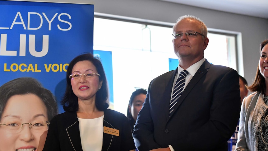 Two politicians wearing black blazers stand next to each other in front of a poster for Gladys Liu.
