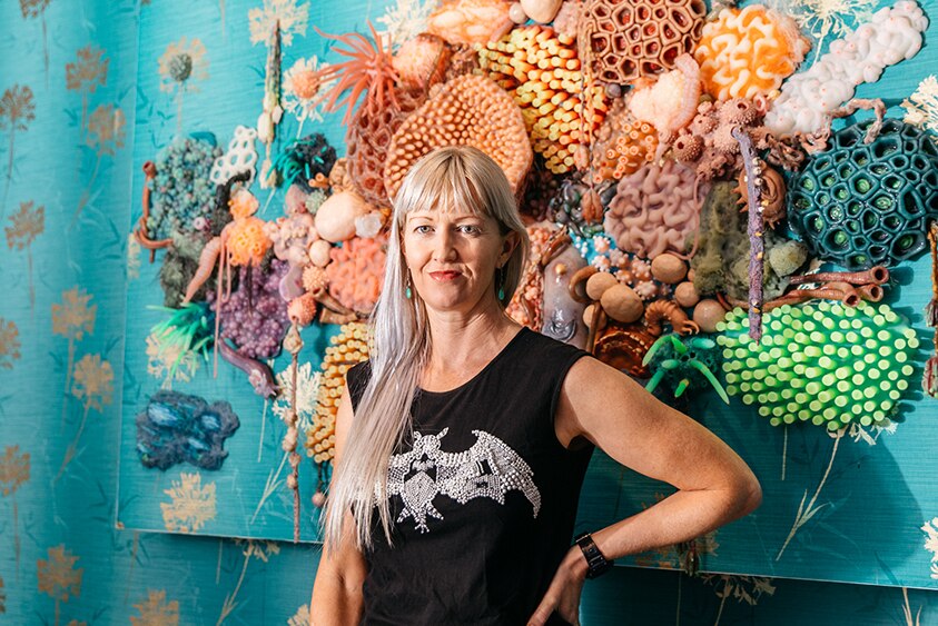 A woman with fringe and long blonde hair stands in front of large coral sculpture fixed to blue wallpapered wall.
