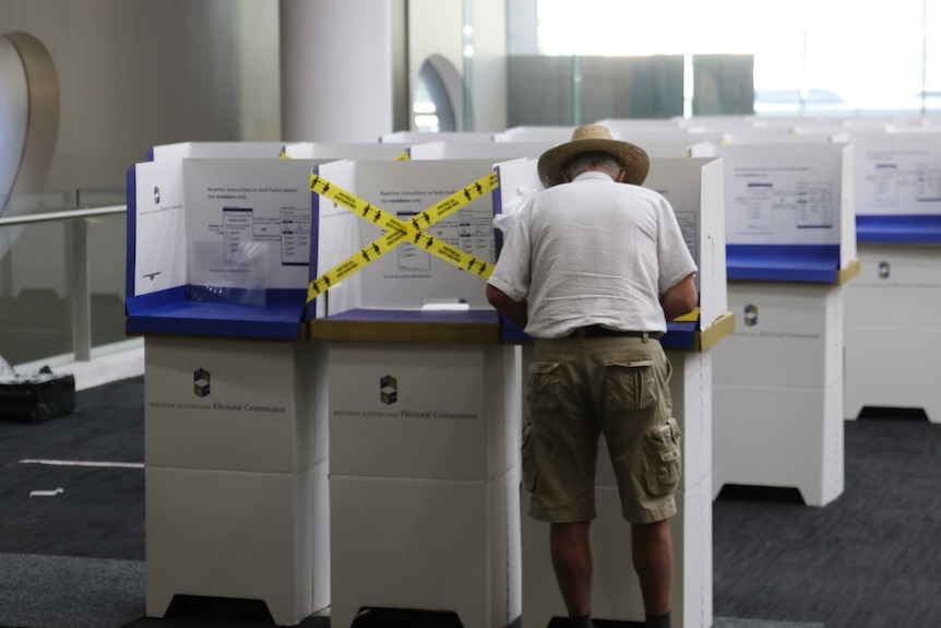 An older man wearing a short sleeve shirt, shorts and a wide brim hat, stands at a polling booth inside a room.