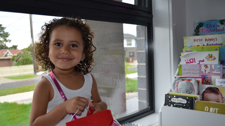 the photo shows a small happy girl surrounded by books holding a bag with a toy in front of a window and smiling at a the camera