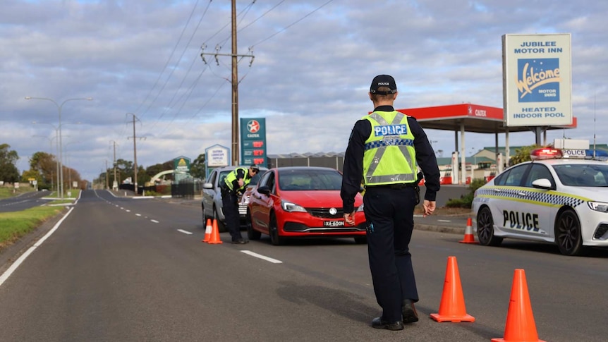 A police officer with his back to the camera walks down a road marked with traffic cones and a queue of cars