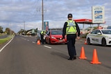 A police officer with his back to the camera walks down a road marked with traffic cones and a queue of cars