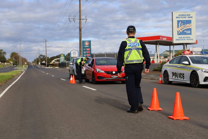 A police officer with his back to the camera walks down a road marked with traffic cones and a queue of cars