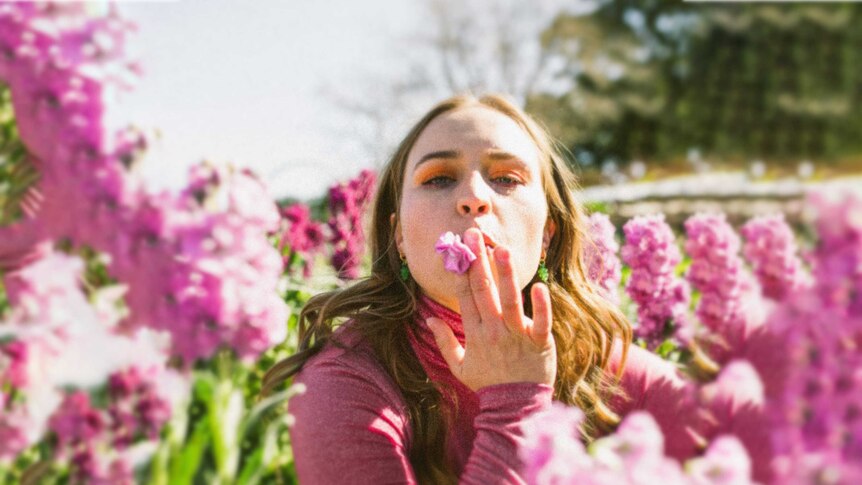 Jack River, wearing a long-sleeve pink turtleneck, crouched in a field of pink flowers, with one flower held at her mouth