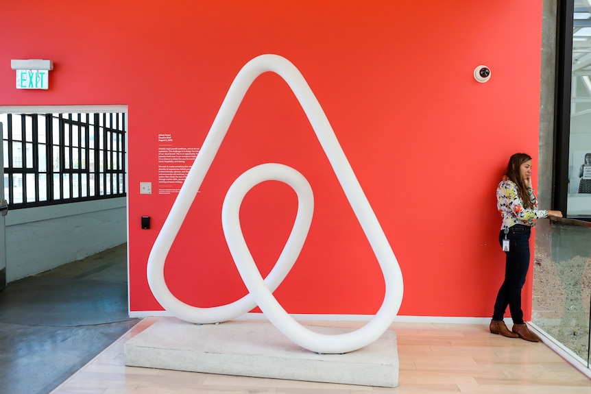 A woman stands and talks on the phone inside an office, next to a large sculpture of the Airbnb logo which is larger than her