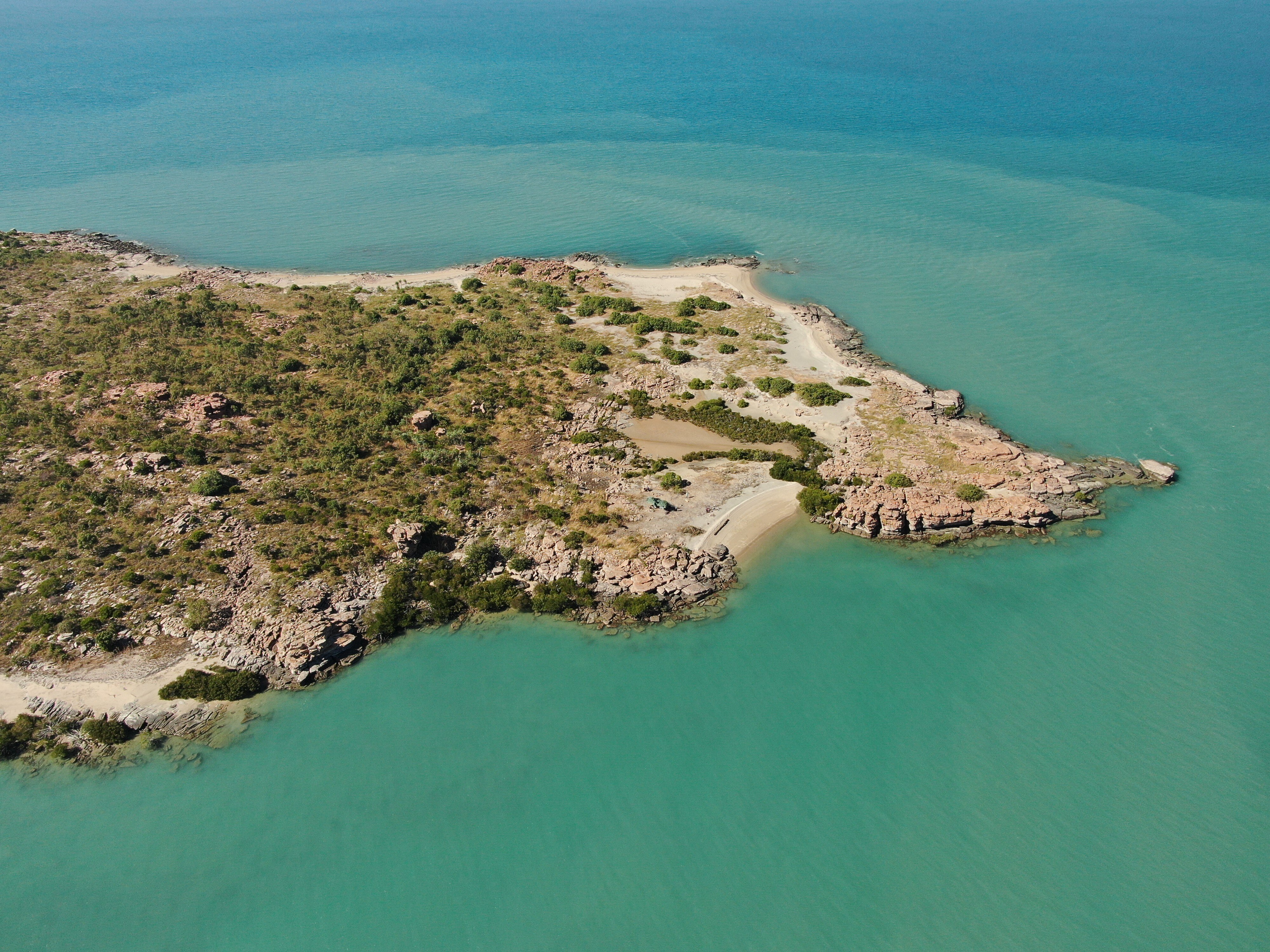 An aerial shot of a coastal island covered in low scrub and surrounded by turquoise sea.