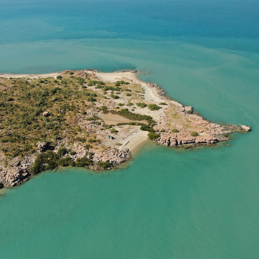 An aerial shot of a coastal island covered in low scrub and surrounded by turquoise sea.