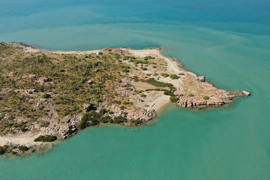 An aerial shot of a coastal island covered in low scrub and surrounded by turquoise sea.