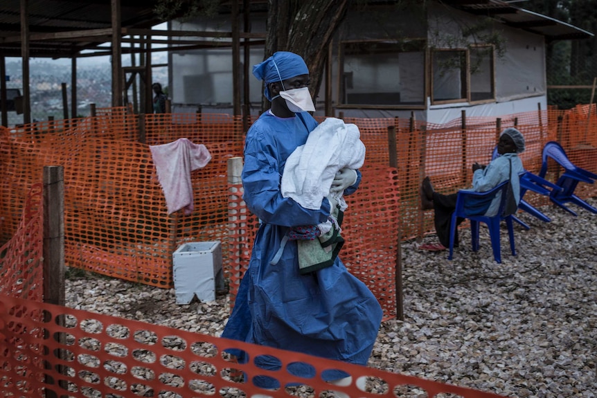 A caretaker dressed in medical scrubs and mask carries a four-day-old baby with suspected Ebola