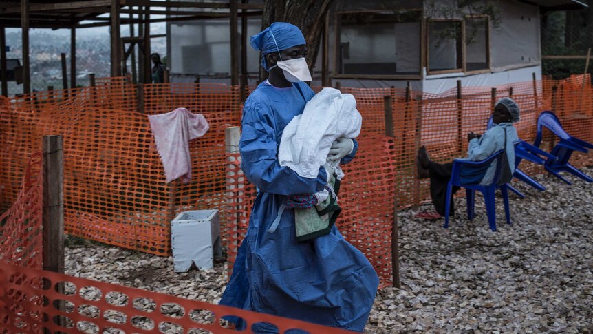 A caretaker dressed in medical scrubs and mask carries a four-day-old baby with suspected Ebola