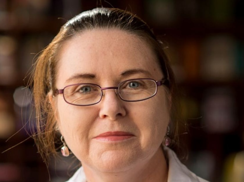 Portrait of Caucasian woman with purple-frame glasses, hair tied back and wearing collared white shirt on blurred background