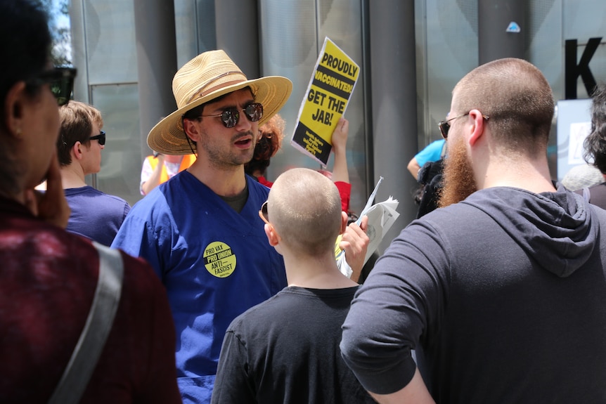 A man wearing sunglasses and a blue t-shirt in a crowd holds up a pamphlet with a pro-vaccination message,