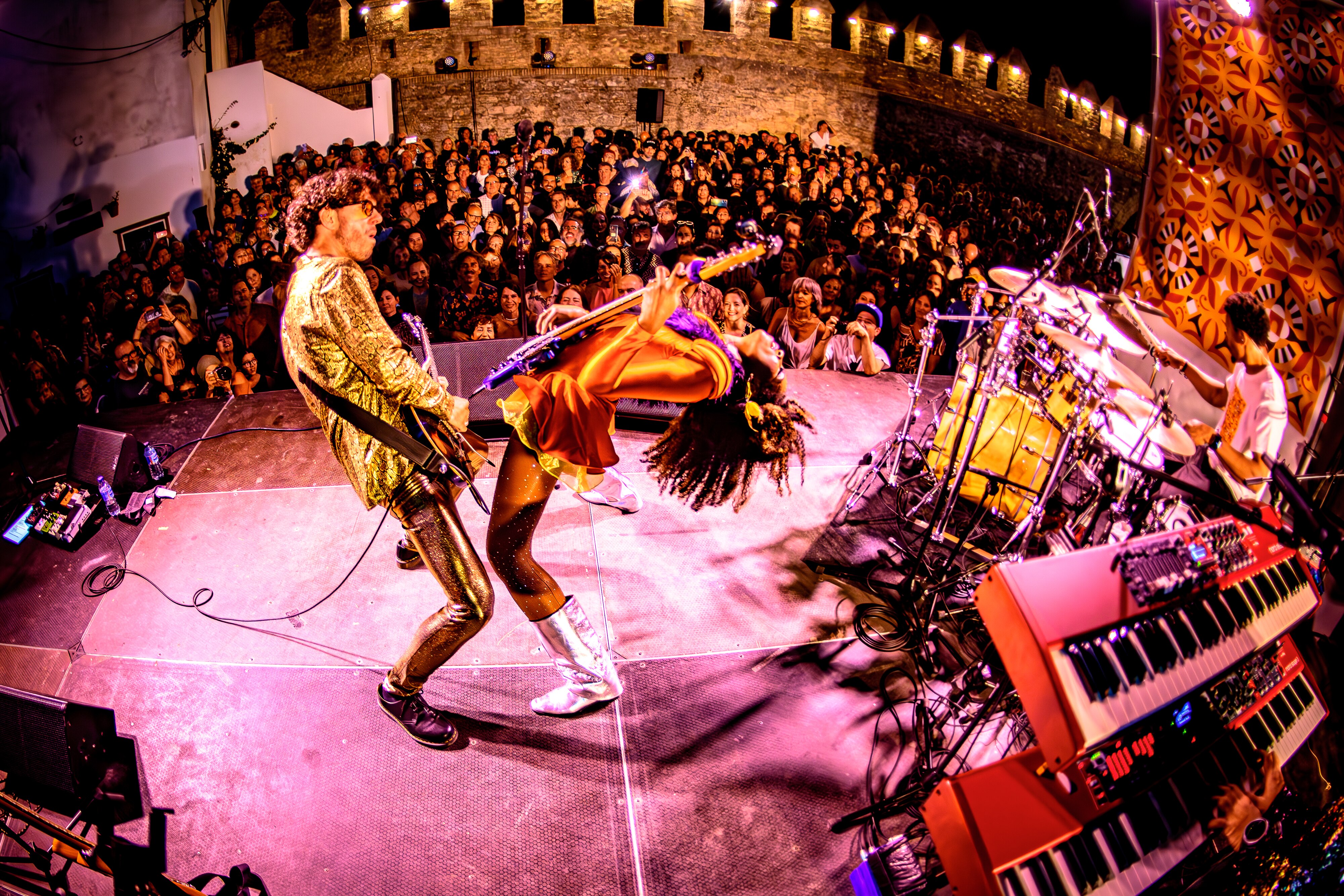 An audience looking at a stage full of musical instruments, a male is playing guitar and a female is leaning back playing bass