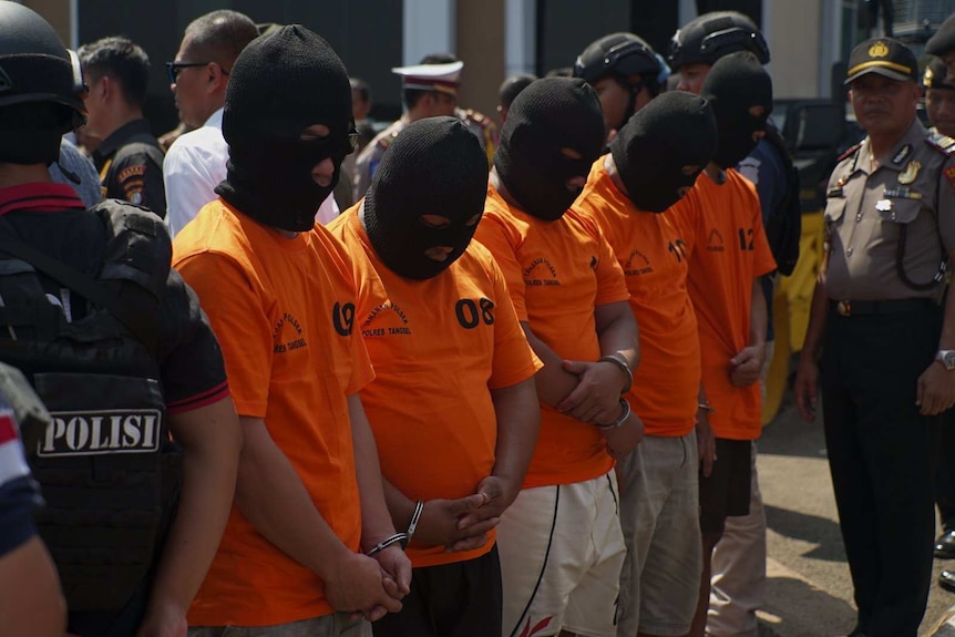 Medium shot of a line of men in orange t-shirts and black balaclavas.