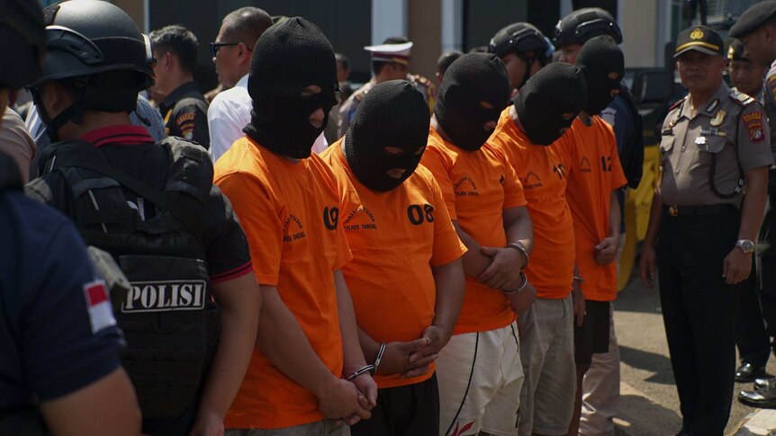 Medium shot of a line of men in orange t-shirts and black balaclavas.