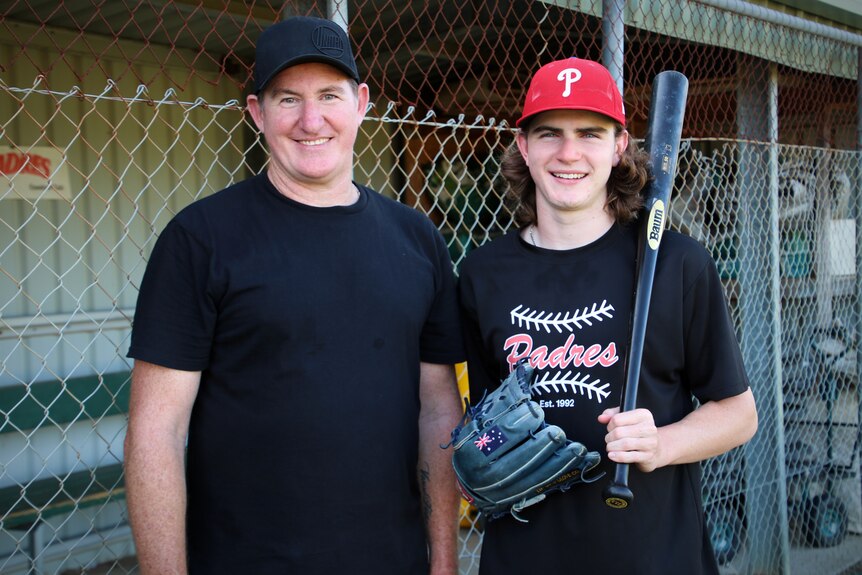 The father and son at a baseball feild. Kai has a bat over his shoulder. 