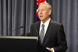 Colin Barnett speaks into microphones at a pedestal in front of a blue curtain and WA flag.