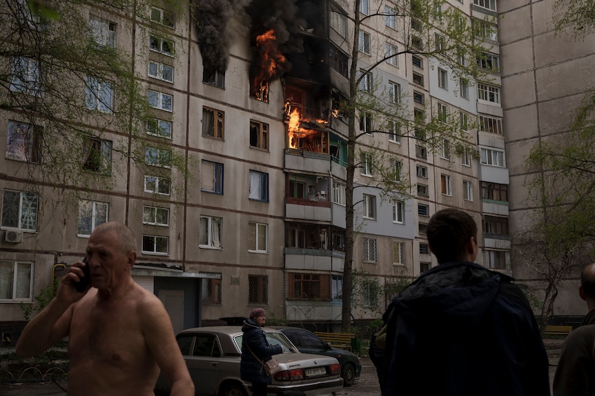 Four people stand outside a large residential building, on which smoke and fire can be seen coming out of a few windows.