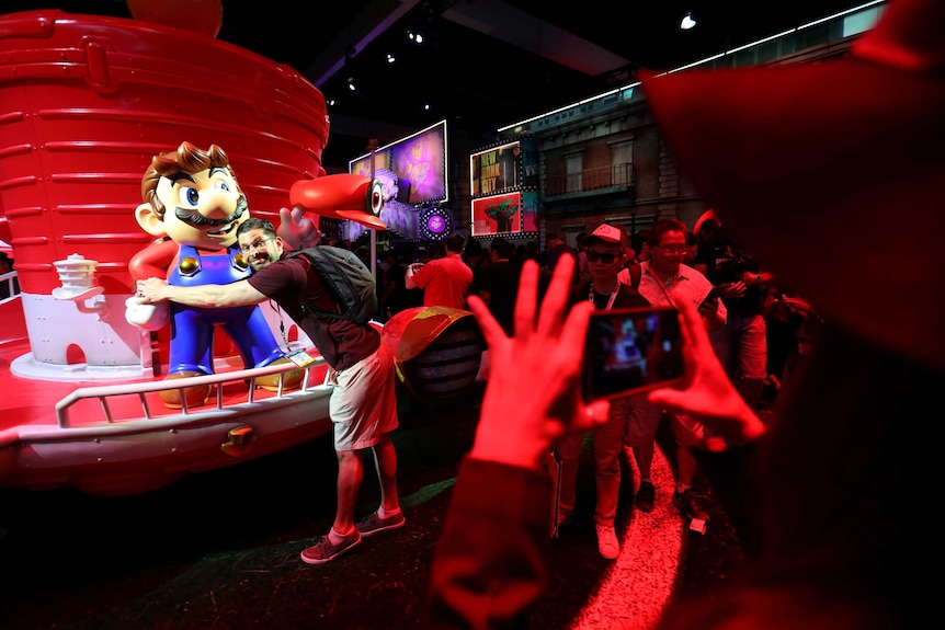 A grown adult man hugs a statue of the Nintendo video character Mario at an E3 video games industry event